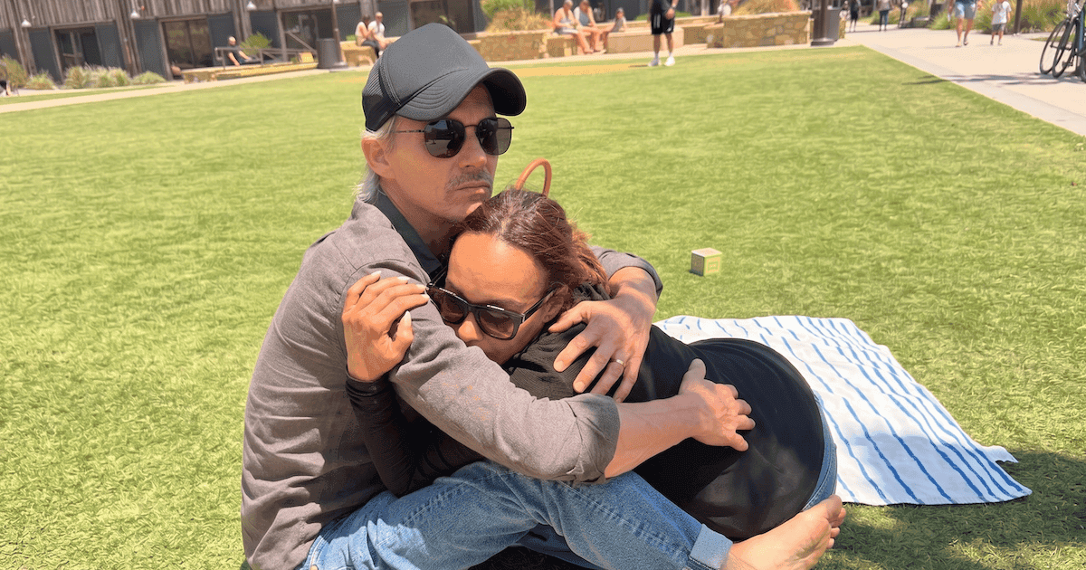 A real couple standing together outdoors — the man's body language reads as quietly present but emotionally contained, which mirrors the article's portrait of a husband who shows up physically while keeping grief locked inside.